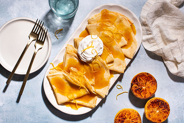 crepes layered on oval platter, topped with butter sauce, orange zest and whipped cream - surrounded by serving plate, forks, water glass and cloth napkin