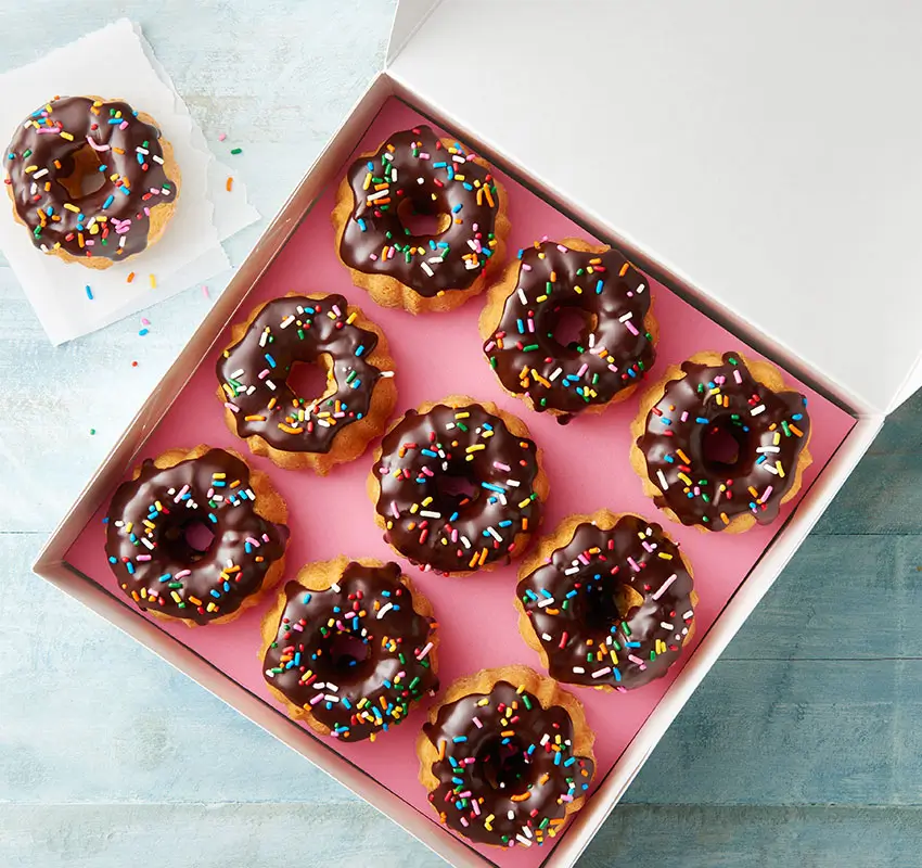 open box of chocolate frosted mini bundt cakes with sprinkles, one "donut" on folded paper towel outside of box