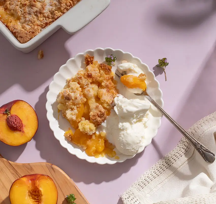 overhead shot of cobbler and scoop of vanilla ice cream on white scalloped plate, surrounded by cobbler in baking dish, fresh peach halves on cutting board and linen napkin