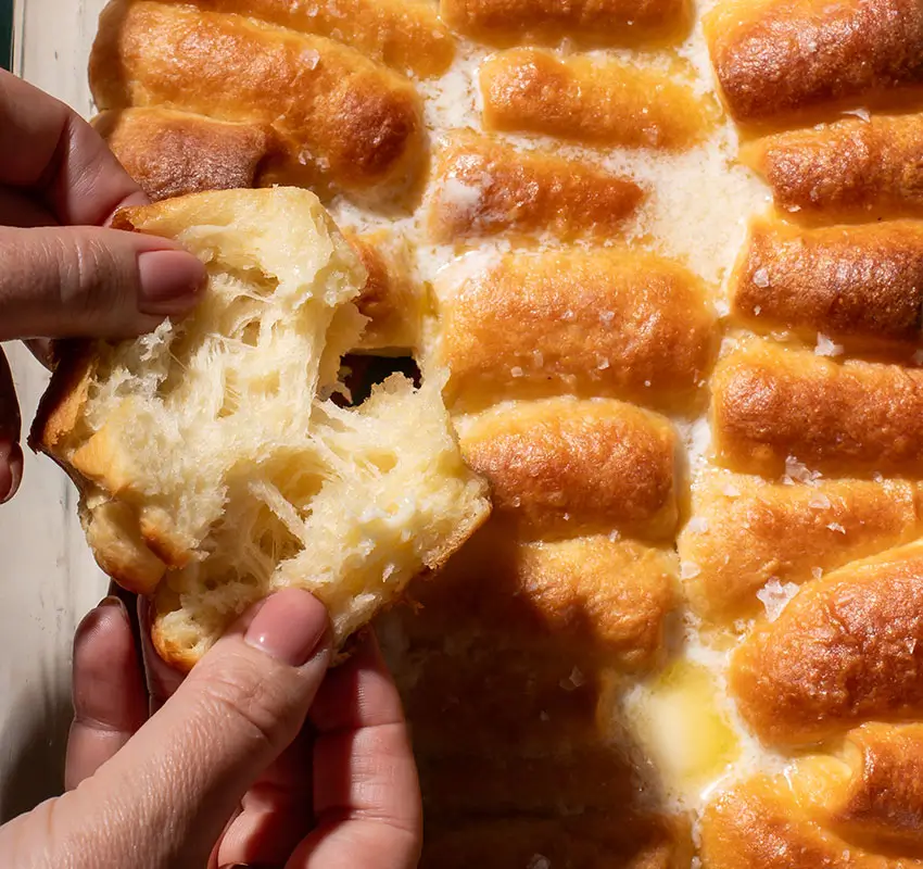 close-up of pan of rolls with melted butter on top, hand pulling apart one roll